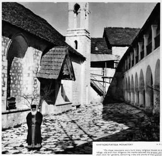 Cobbled courtyard of Khrysoroyiatissa Monastery with a bell tower, arched gallery and a man in dark robes holding a staff