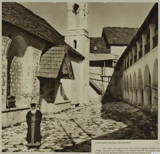 Courtyard of Khrysoroyiatissa Monastery with a stone bell tower, an arcaded gallery and a bearded man holding a staff