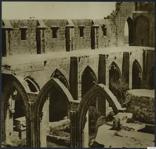 Ruined monastery building with a row of pointed arches and windowed walls, with bushes and fallen stone blocks in the foreground