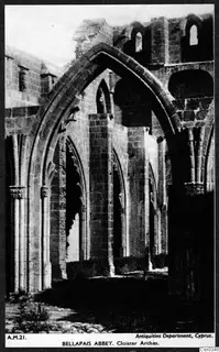Pointed arches and stone piers of the cloister at Bellapais Abbey, with ruined walls rising behind.