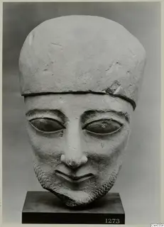 Limestone head of a bearded figure with a tall flat-topped cap, standing on a dark square base.