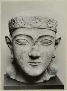 Limestone head of a figure with a flat-topped headband decorated with rosettes, shown on a dark stand against a grey backdrop.
