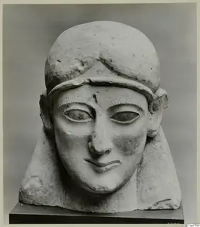 Limestone sculpted head of a figure wearing a close-fitting cap, with a smiling mouth and almond-shaped eyes.