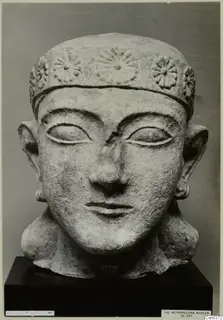 Frontal view of a carved limestone head with a rosette-decorated headband and large ears, on a black stand