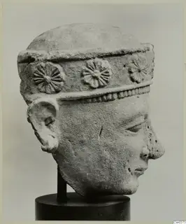 Carved limestone head of a figure wearing a band with rosette ornaments, shown on a dark stand