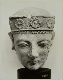 Limestone head of a figure wearing a band with rosettes, on a black stand with a number label.