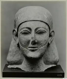 Carved limestone head with a close-fitting headdress and broad ears, nose broken, set on a dark wooden base