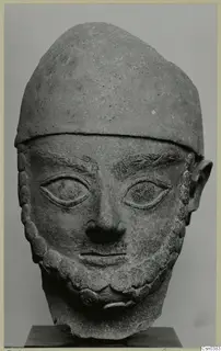 Carved stone head of a bearded man in a close-fitting cap, shown frontally on a dark stand.