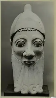 Limestone sculpture of a head with a tall rounded headdress, wide almond-shaped eyes and a broad nose, standing on a dark plinth