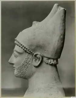 Limestone sculpture of a bearded head in profile, facing left, wearing a tall headdress with a curled neck band.