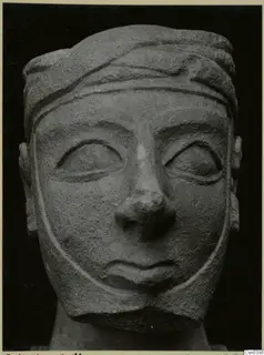 Carved limestone head with a banded headdress and broad face, seen from the front against a dark background