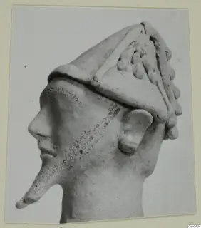 Terracotta head in profile with a tall conical headdress, applied ridges and knobs, and a dotted pattern on the cheek