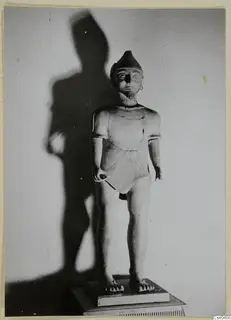 Standing terracotta statue of a figure in a pointed cap and short-sleeved tunic, set on a stepped base against a wall.