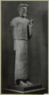 Standing male terracotta statue with a headband, beard and raised right hand, shown on a dark plinth against a grey backdrop.