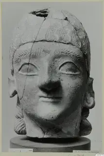 Terracotta head with incised curled hair and a broken crown, shown on a grey cylindrical stand