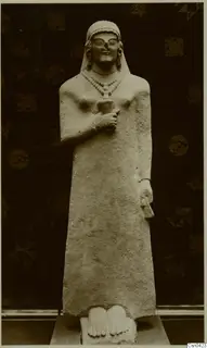 Limestone standing statue of a figure with a close-fitting head covering and beaded necklace, photographed against a dark patterned cloth.