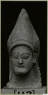 Limestone head statue with a tall conical headdress and closed eyes, shown against a black background.