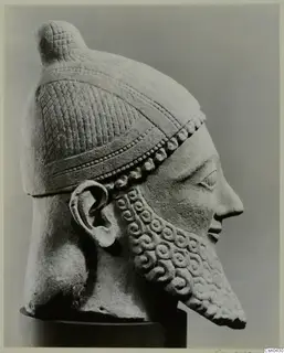 Limestone head of a bearded male figure in a helmet with a knob crest, shown in profile to the right.