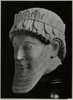 Limestone head of a statue with a leafy or petal-patterned headdress and a long, textured beard.
