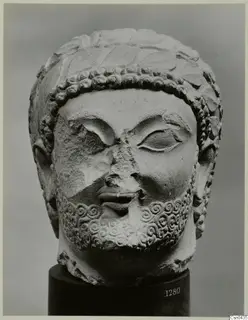 Limestone sculpture head of a bearded man, with curled hair and beard; the nose and part of the face are broken.