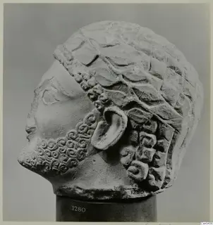Limestone head sculpture in left profile, with a curled beard and hair in overlapping locks above a band.