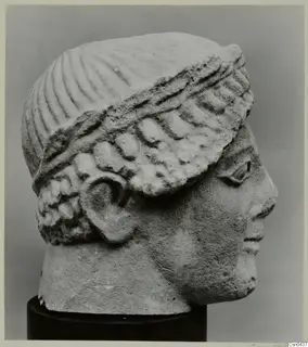 Limestone head of a figure in profile, with a ribbed headdress and curled hair, on a dark stand