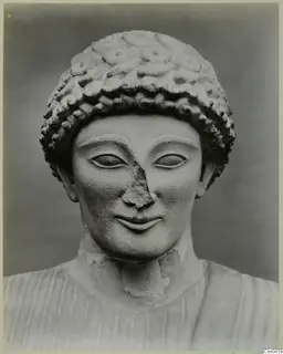 Limestone head of a statue with curled hair in a cap of curls and a smiling face, nose chipped.