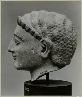 Limestone head of a figure in profile, facing left, with ridged hair and a carved ear, on a dark wooden stand.