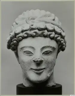 Limestone head of a figure with curled hair in tiers, mounted on a dark pedestal against a grey backdrop.