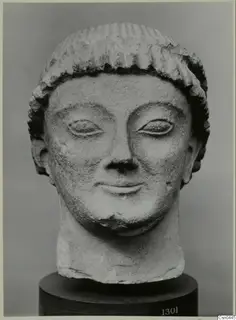 Limestone head of a figure with grooved hair, eyes and smiling mouth, shown on a dark cylindrical base.