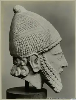 Limestone head of a bearded figure in a tall, grid-patterned helmet, shown in profile on a dark stand.