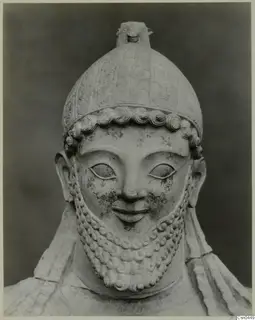 Limestone sculpture of a bearded male head in a helmet with a crest, photographed against a dark backdrop.