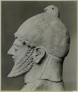 Limestone sculpture of a bearded head in profile, wearing a helmet with incised decoration and a bird-like crest.