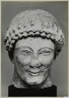 Limestone head of a figure with a curled hair band, photographed against a grey backdrop on a black stand.