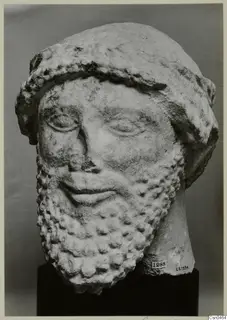Limestone head of a bearded male sculpture with a wide band over the hair, shown against a grey backdrop.
