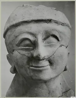 Terracotta head of a figure with a flat cap, large heavy-lidded eyes and a broad smile, cracked across the face