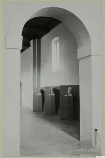 Three sculpted heads on dark pedestals in a museum gallery, seen through a tall arched doorway.