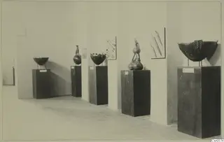 Five objects on pedestals in a gallery: bowls, a tall-necked flask, a stacked group of vessels, and blade-like tools on the wall