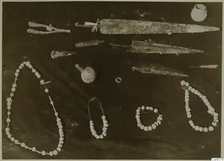 Metal blades, a small vessel, a ring and four strings of white beads laid out on a dark cloth