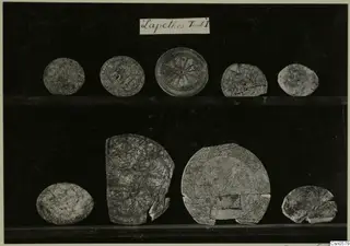 Nine round objects from Lapithos, Aj. Anastasia, T.I, some with patterns, shown on two shelves against a dark background.