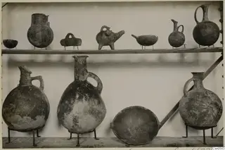 Two wooden shelves of pottery vessels on iron stands, mostly jugs and juglets, with a few bowls.