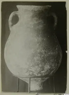 Two-handled pottery vessel with a wide rim, standing on a three-rod metal stand against a dark background.
