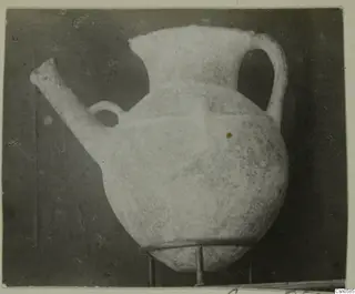 Pale pottery jug with a long spout, a small loop handle and a tall strap handle, shown on a metal stand