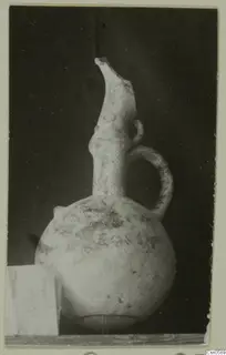 Pottery vessel with a globular body, tall flaring spout and strap handle, photographed on a wooden shelf