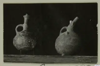 Two pottery juglets with loop handles, one with a tall neck and one with a short upward spout, on a wooden shelf