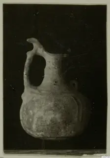 Pottery jug with a strap handle and pinched spout, photographed against a dark background