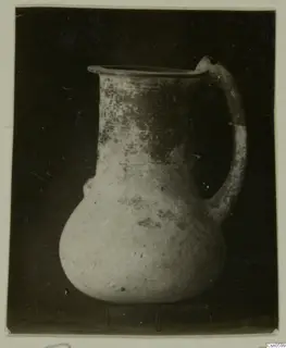 Pale pot with a flared rim and strap handle, with dark patches on the upper body, against a dark background.