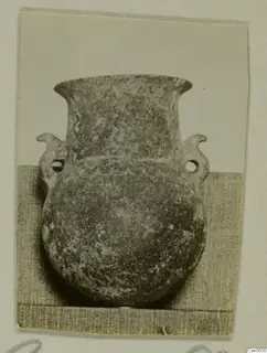 Round-bodied pot with a flared neck, two perforated handles and wavy lines on the neck, photographed on a grey cloth
