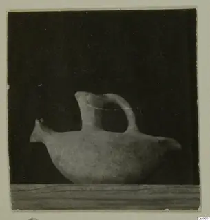 Pale pottery vessel with a beak-like spout and a tail-like projection, shown in side view on a wooden ledge