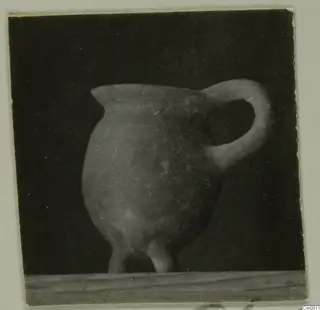 Rounded pot with a pinched lip and a loop handle, with short feet, standing on a wooden board against a dark background.